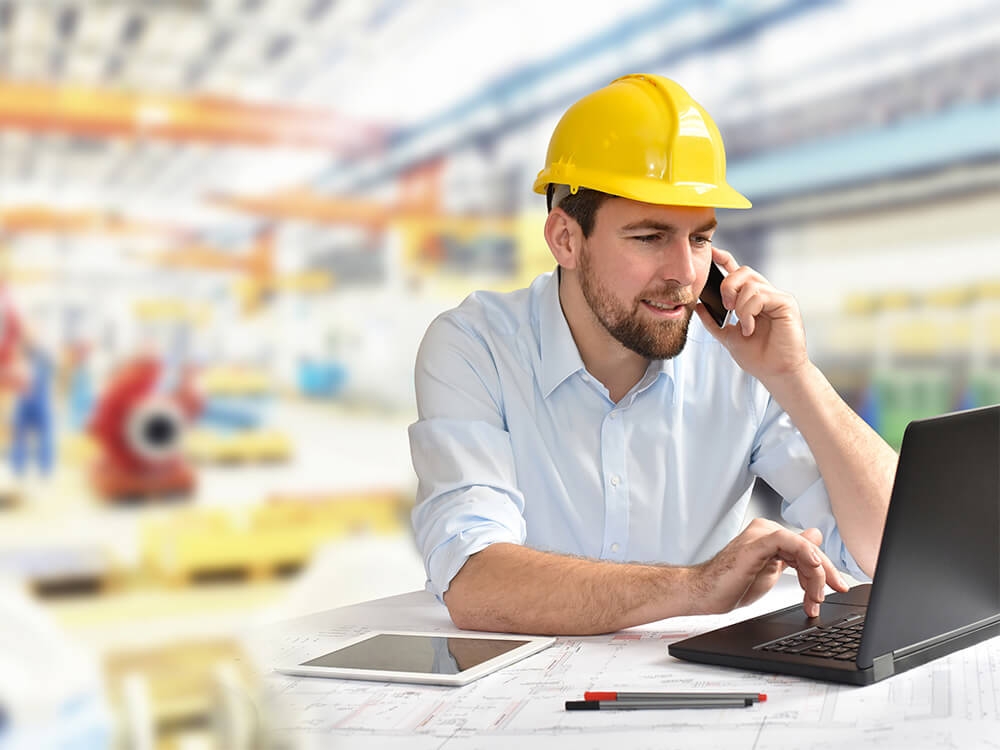 Ein telefonierender Arbeiter mit einem gelben Schutzhelm, mit einem Laptop und einem Tablet auf einem Schreibtisch in einer geschäftigen Industrieumgebung.